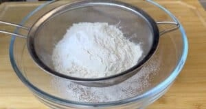 Sifting flour through a fine mesh sieve into a glass bowl on a wooden surface for baking preparation.